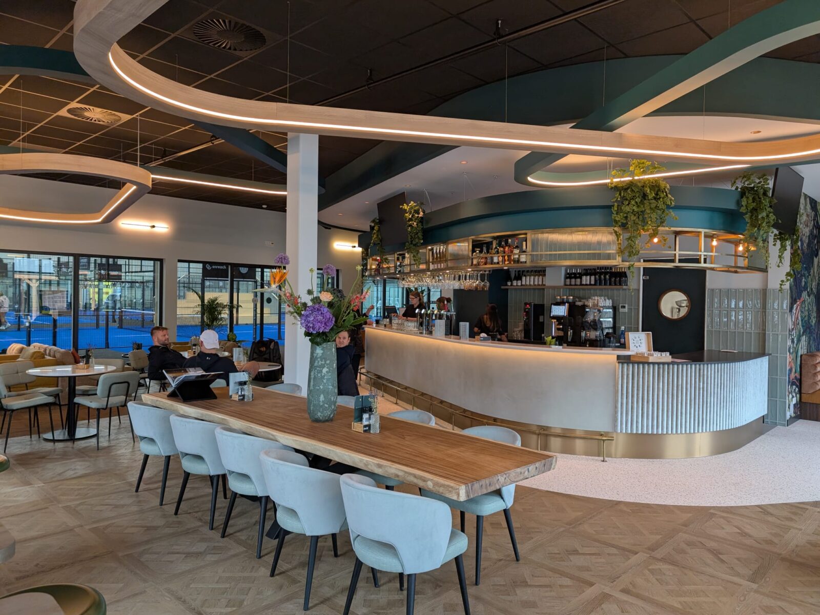 The hospitality area of Poort Padel with a round bar, a long wooden table and a view of the blue padel courts
