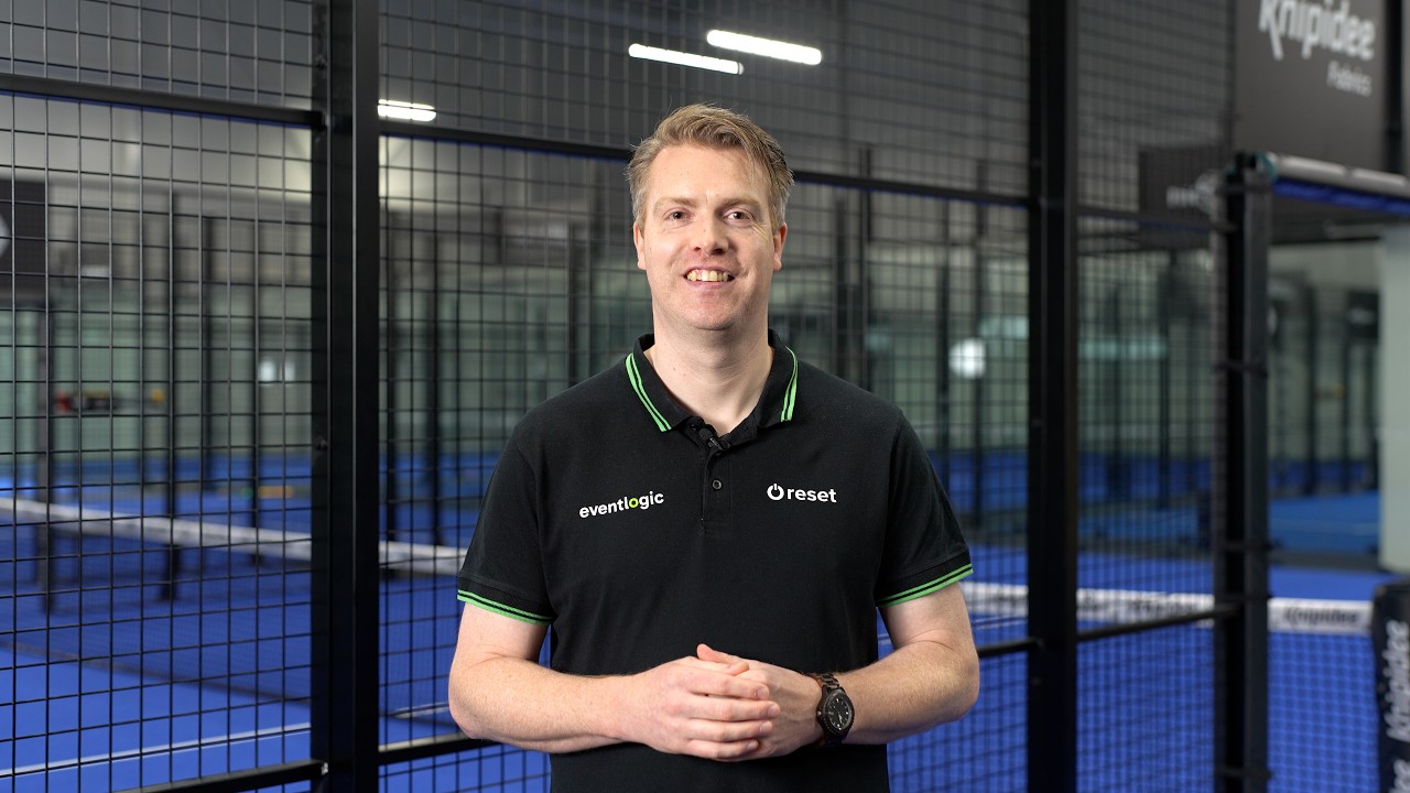 The video presenter in a black Eventlogic and Reset shirt in front of a padel court.